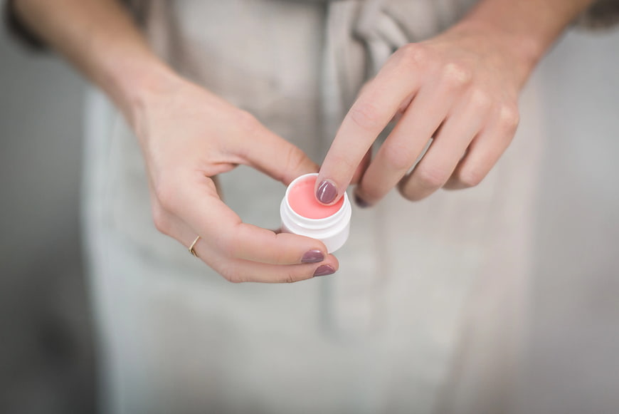 Woman applying pink lip balm from small white jar