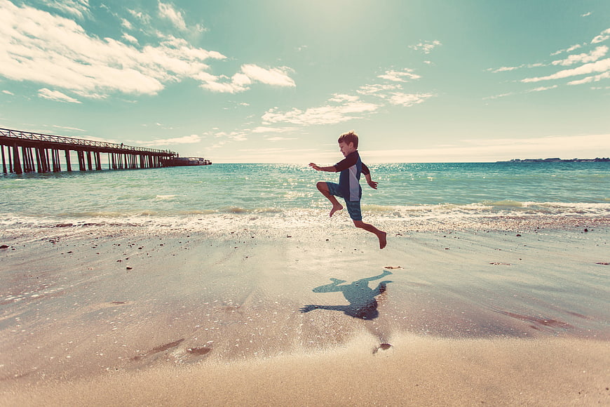 Young boy jumping on a sunny beach with a pier in the background