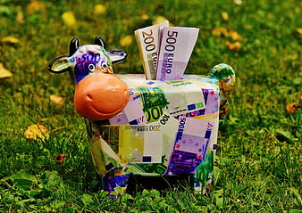 Cow-shaped piggy bank covered in euro banknotes on grass