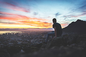 Man sitting on cliff overlooking city at sunset