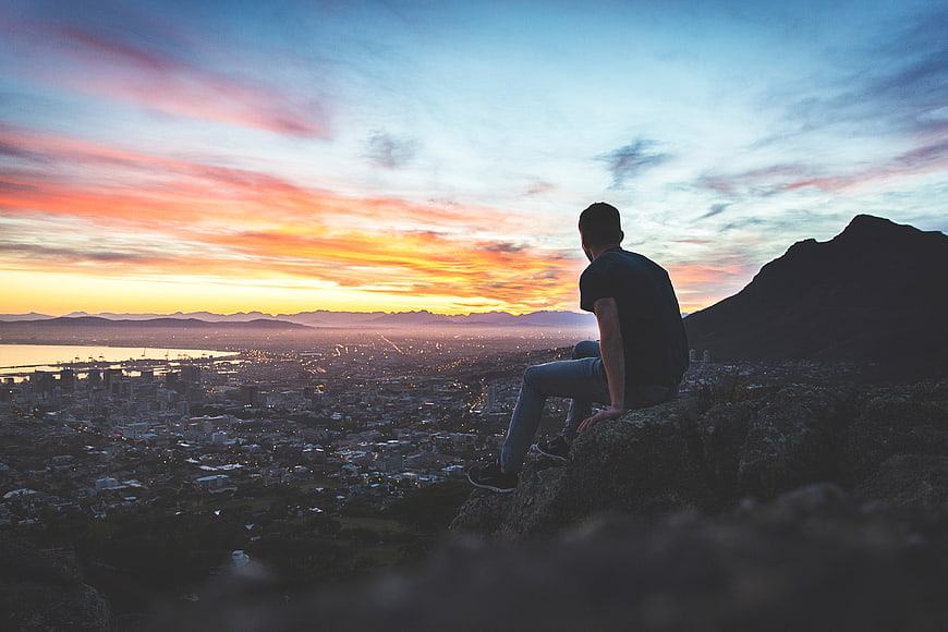 Man sitting on cliff overlooking city at sunset