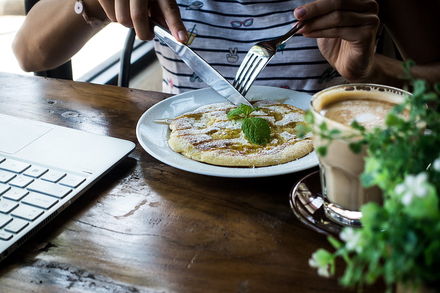 Woman eating honey pancake with coffee and laptop