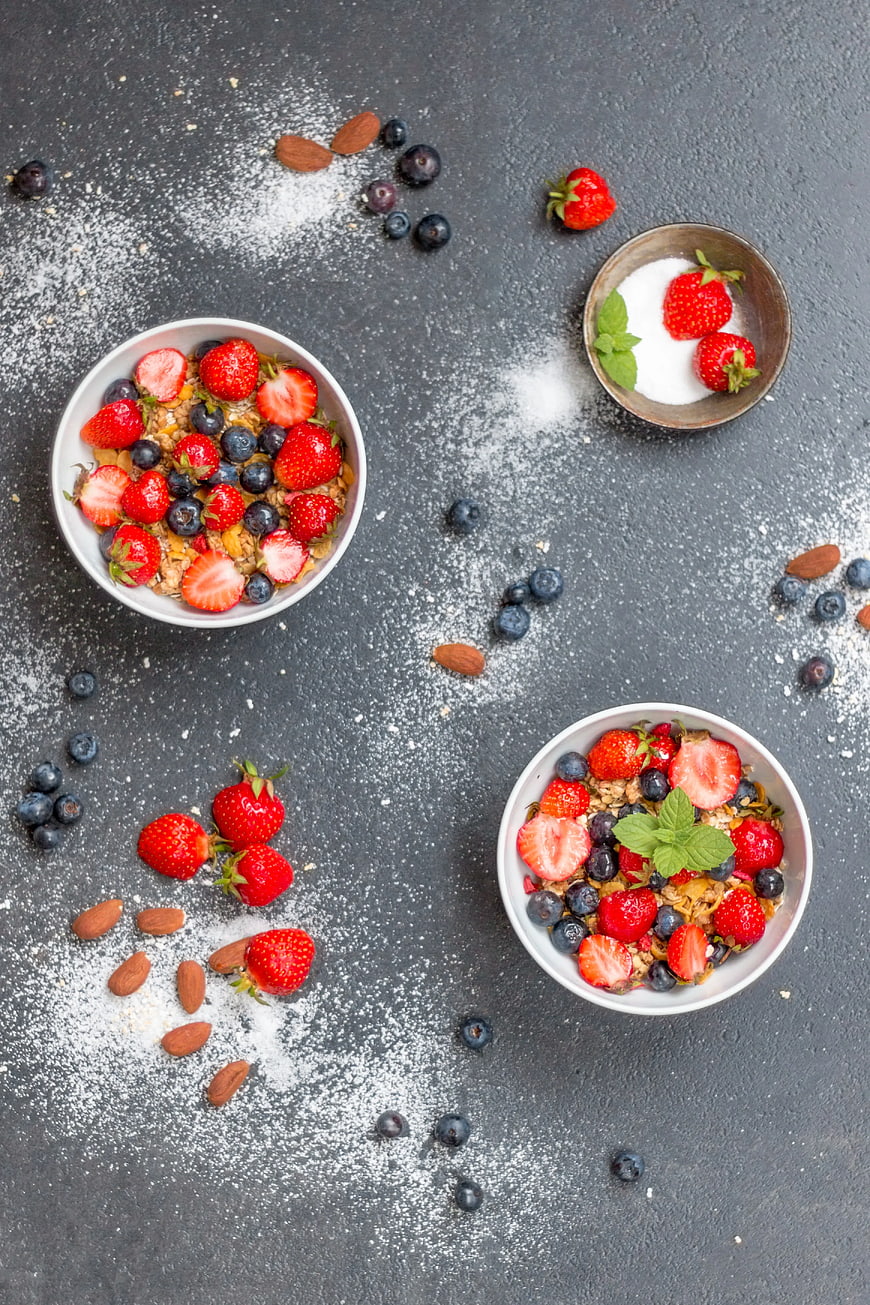 Overhead view of healthy breakfast bowls with granola, strawberries, and blueberries on a dark textured background.