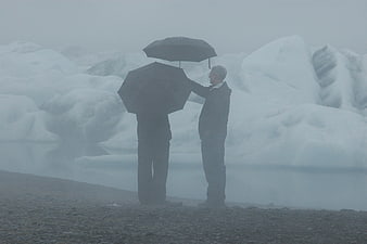 Two men with umbrellas standing by a foggy glacial lake