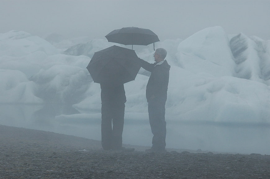 Two men with umbrellas standing by a foggy glacial lake