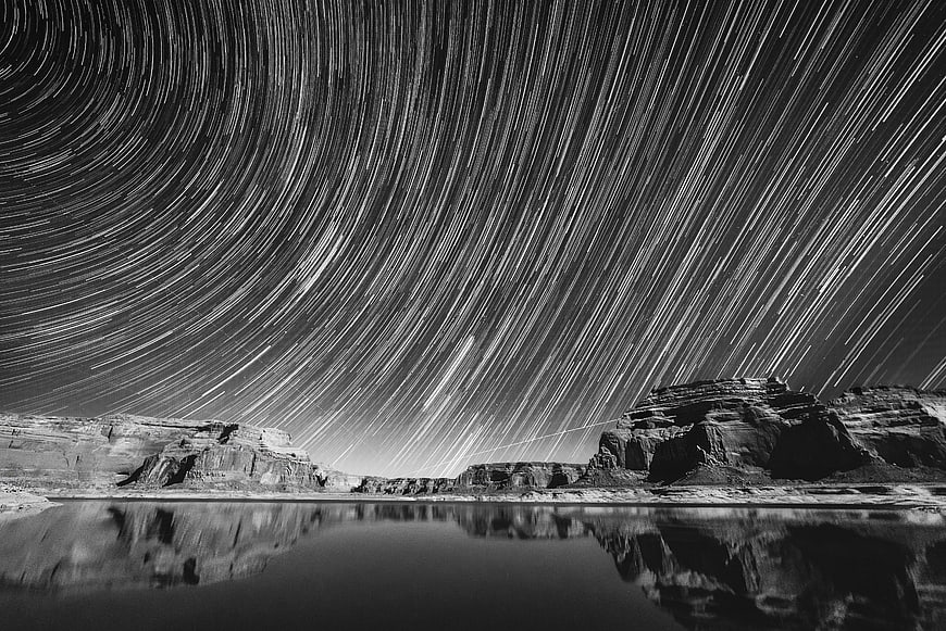 Black and white star trails over desert canyon with water reflection