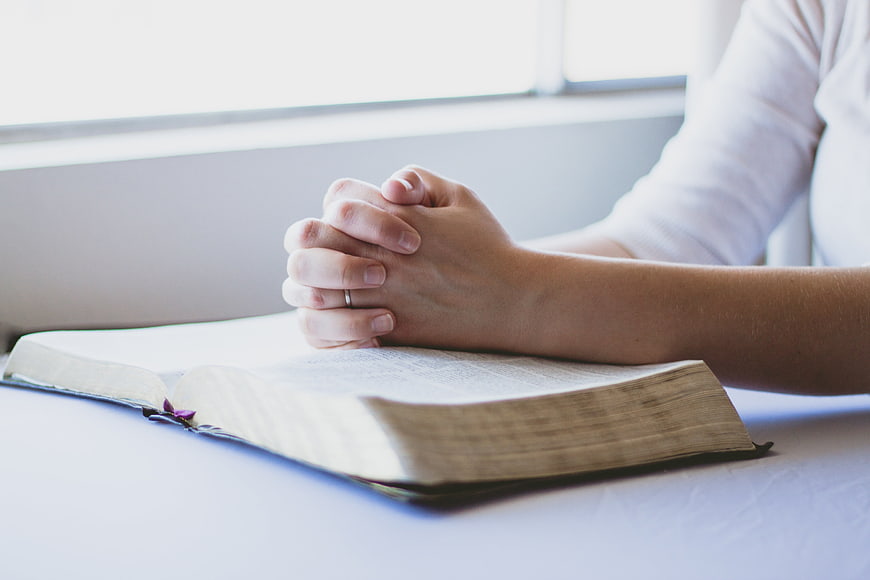 Praying Hands Resting on Open Bible by Window