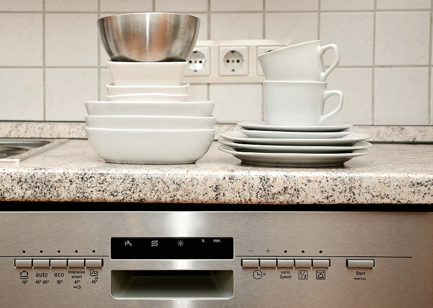 Stacked clean white dishes on granite countertop above dishwasher