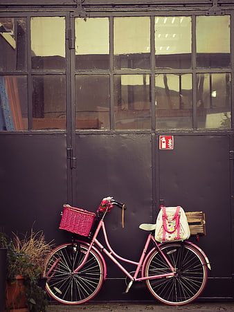 Pink vintage bicycle with basket and bag parked against dark industrial wall