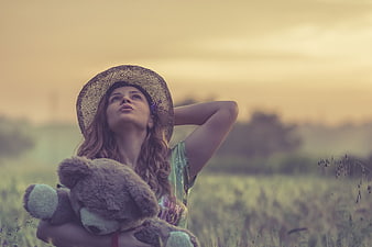 Woman in straw hat holding teddy bear in field at sunset