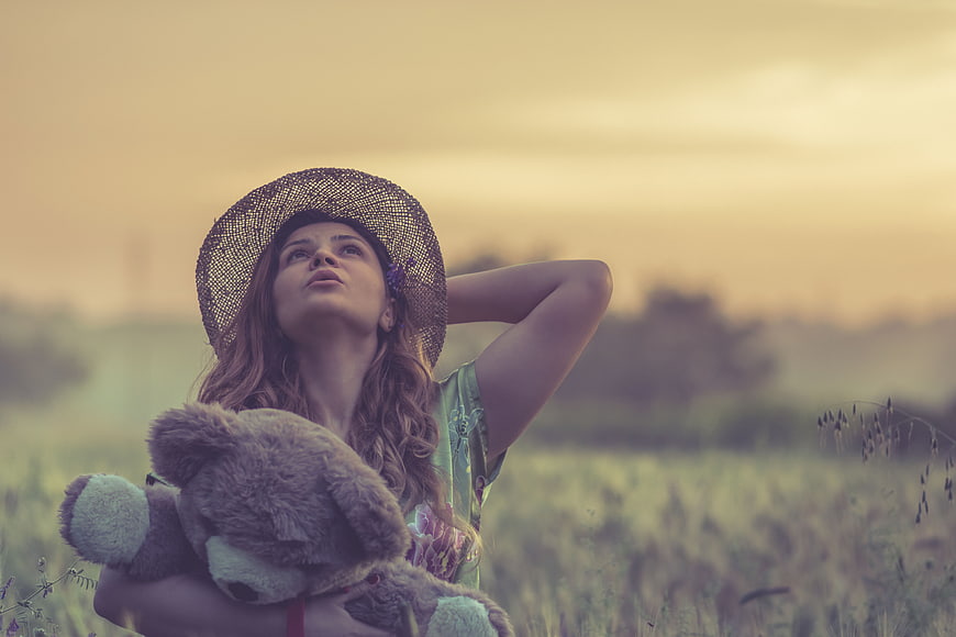 Woman in straw hat holding teddy bear in field at sunset