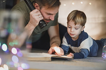 Father and son reading book with flashlight at night