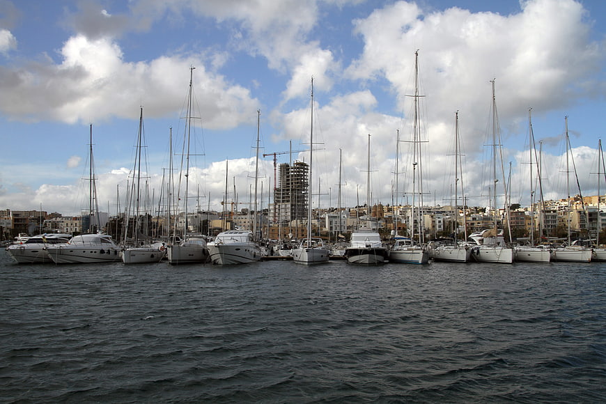 Sailboats docked in a marina with city skyline and clouds