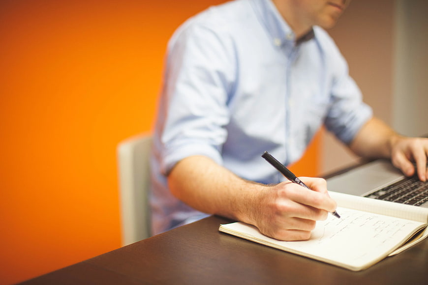 Man Writing in Notebook at Desk with Laptop