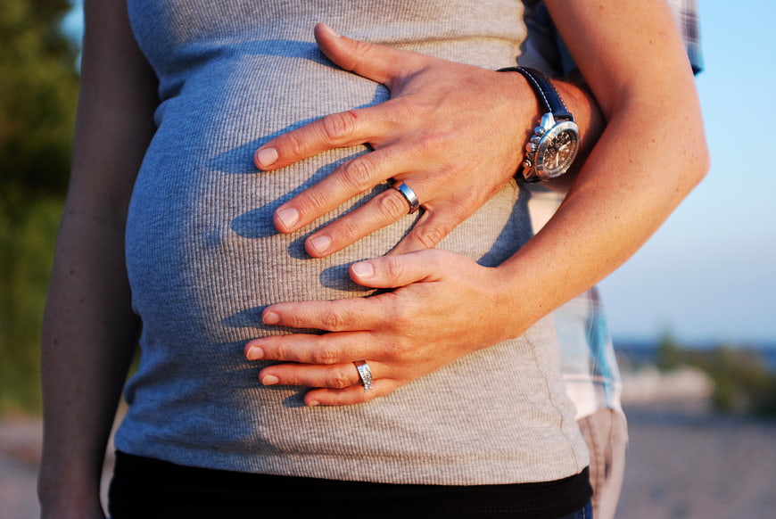 Pregnant woman holding belly with partner's hands outdoors