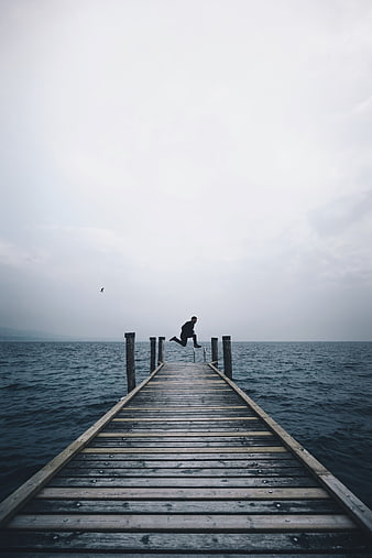 Man jumping off wooden pier into ocean on overcast day