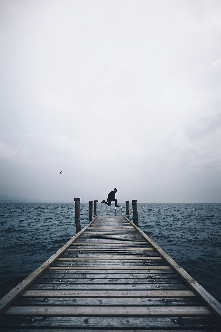 Man jumping off wooden pier into ocean on overcast day