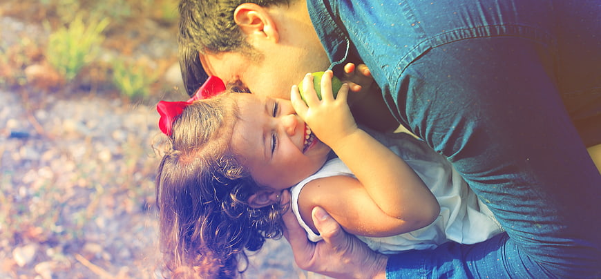 Father kissing laughing daughter with green apple outdoors