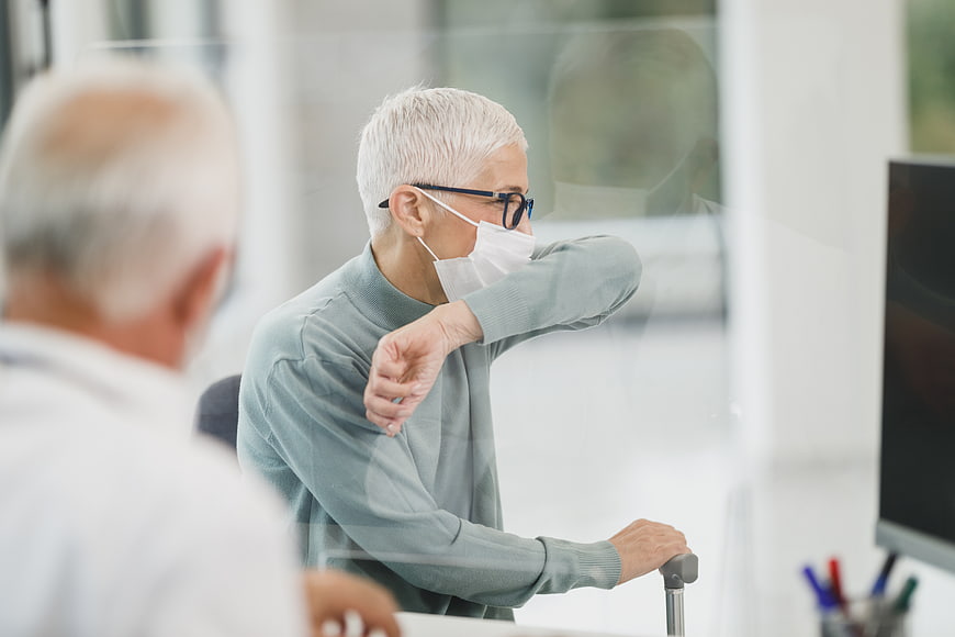 Senior woman with crutch coughing into mask at clinic