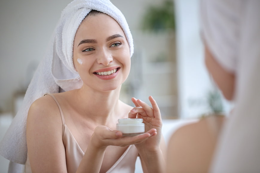Happy woman applying face cream in bathroom mirror