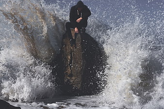 Man in black hoodie sitting on rock amidst crashing ocean waves