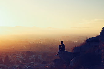 Silhouette of man sitting on cliff edge overlooking city at sunset