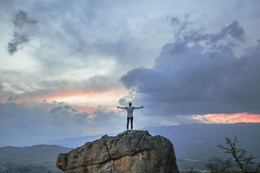 Man standing on rocky peak with arms outstretched at sunset