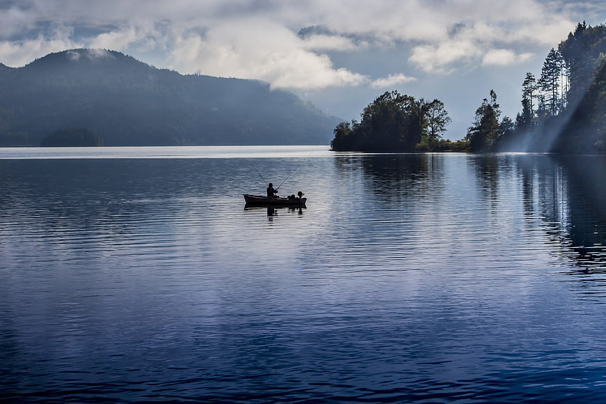 Fisherman in small boat on calm lake with mountain reflections