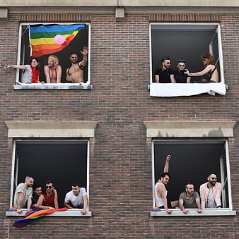 LGBTQIA+ men waving rainbow pride flags from brick building windows