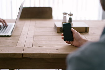 Person Using Smartphone and Laptop at Wooden Desk