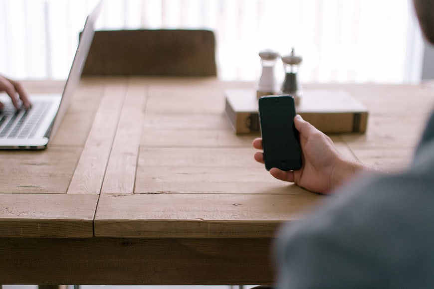 Person Using Smartphone and Laptop at Wooden Desk