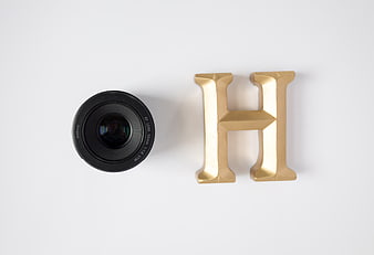 Black Camera Lens and Gold Letter H on White Background