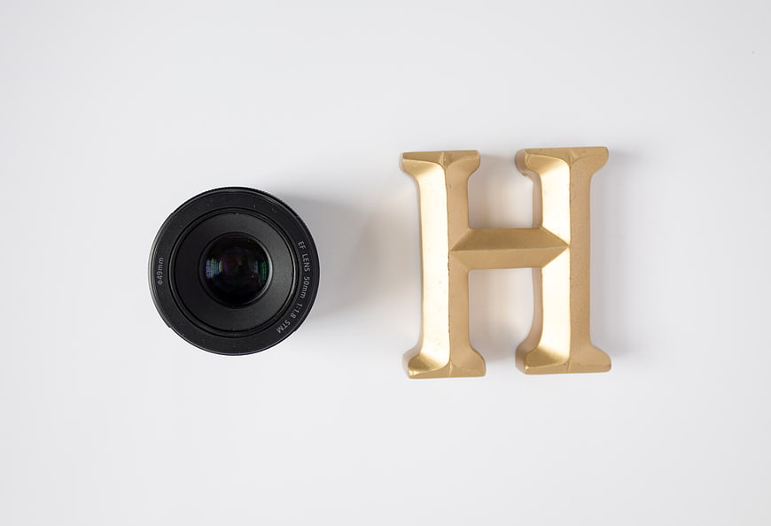 Black Camera Lens and Gold Letter H on White Background