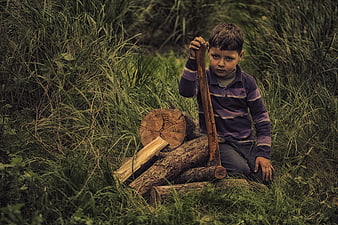 Young boy holding axe with chopped wood in grassy field