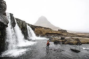 Hiker in Orange Jacket Standing by Waterfall with Kirkjufell Mountain