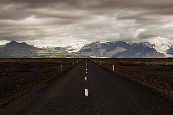 Empty asphalt road leading to snow-capped mountains under cloudy sky