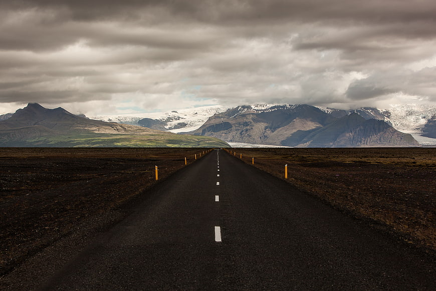 Empty asphalt road leading to snow-capped mountains under cloudy sky