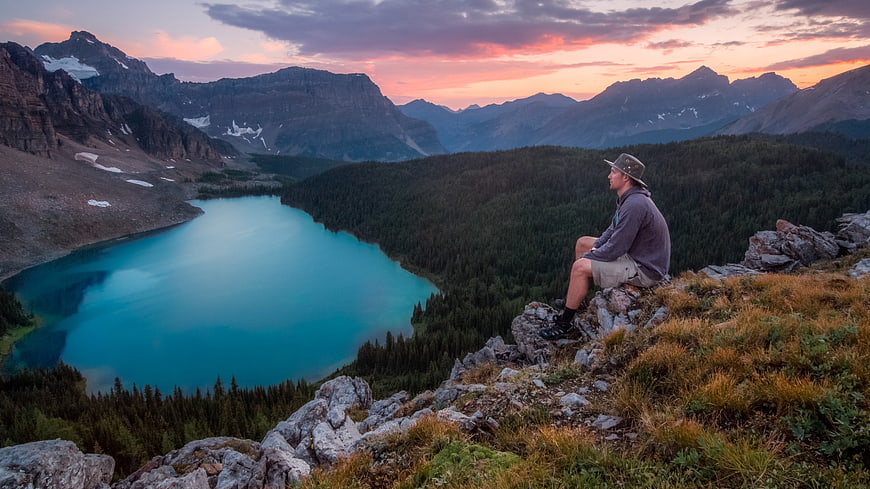 Hiker sitting on rocky mountain peak overlooking turquoise alpine lake at sunset