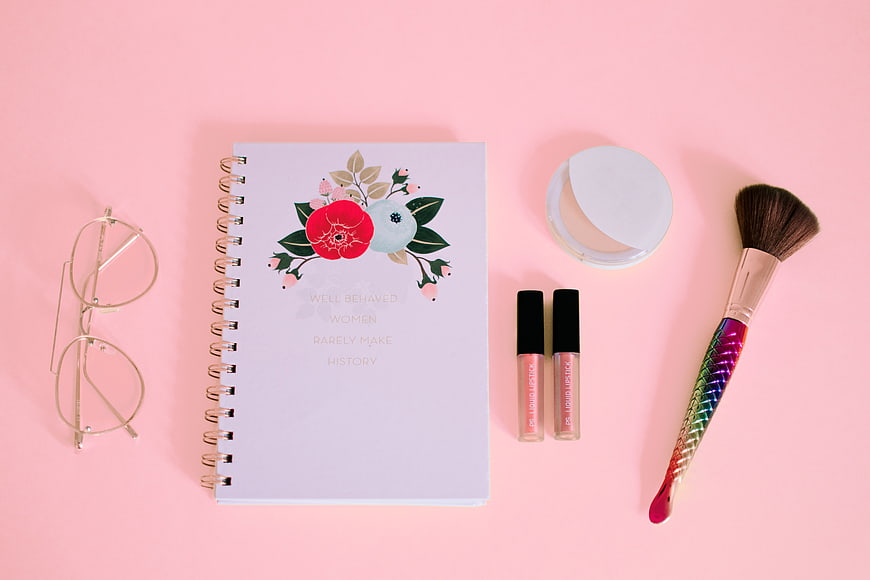 Flat lay of makeup, notebook, and glasses on pink background