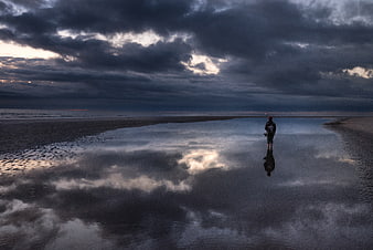 Lone figure standing in tidal pool reflecting stormy sky