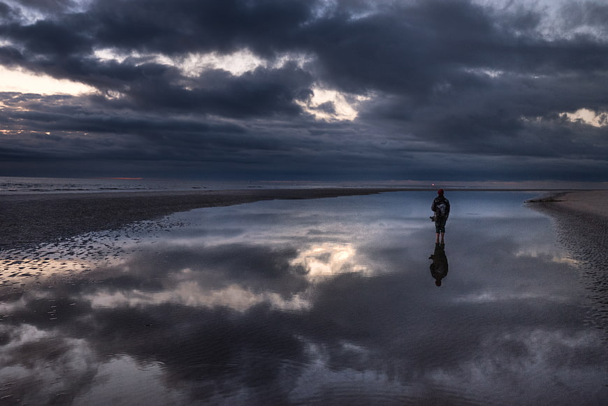 Lone figure standing in tidal pool reflecting stormy sky
