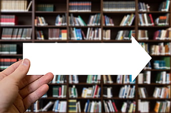 Hand holding white arrow sign in front of blurred library bookshelves