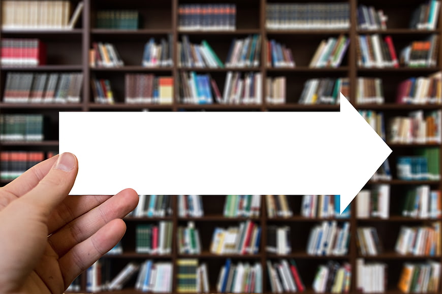 Hand holding white arrow sign in front of blurred library bookshelves