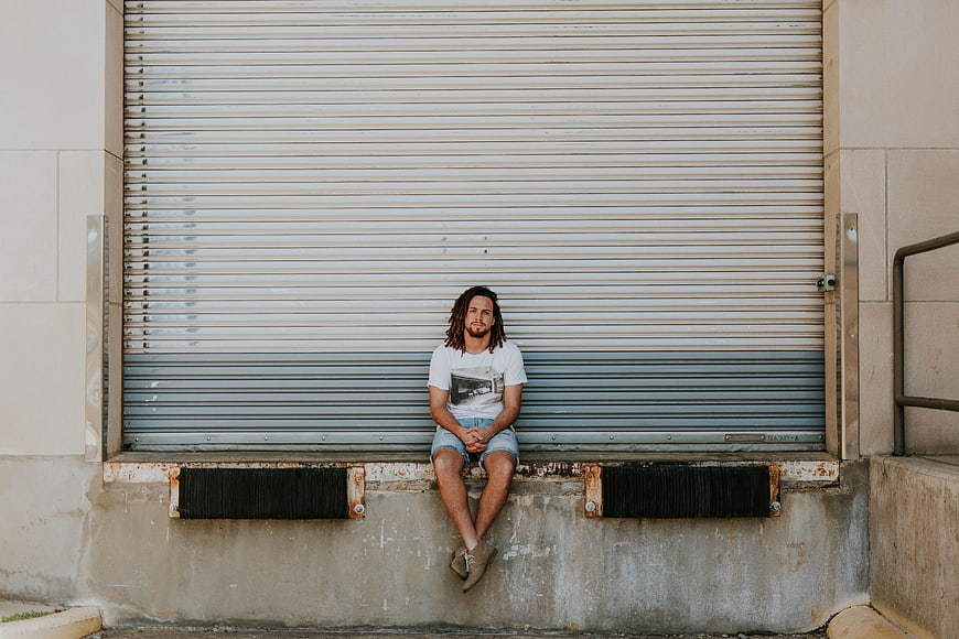 Man with dreadlocks sitting on concrete ledge in front of metal shutter