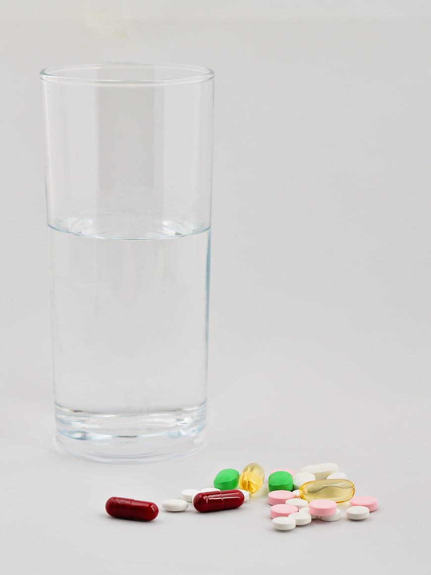 Glass of water with scattered colorful pills and capsules on white background