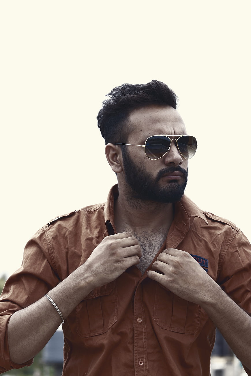 Bearded man in brown shirt and aviator sunglasses adjusting collar