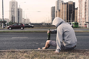 Lonely man in hoodie sitting on roadside curb with beer bottle