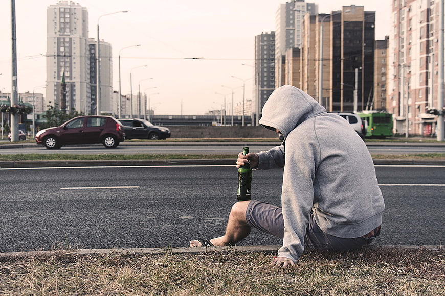 Lonely man in hoodie sitting on roadside curb with beer bottle