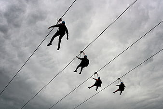 Silhouettes of people zip lining against cloudy sky