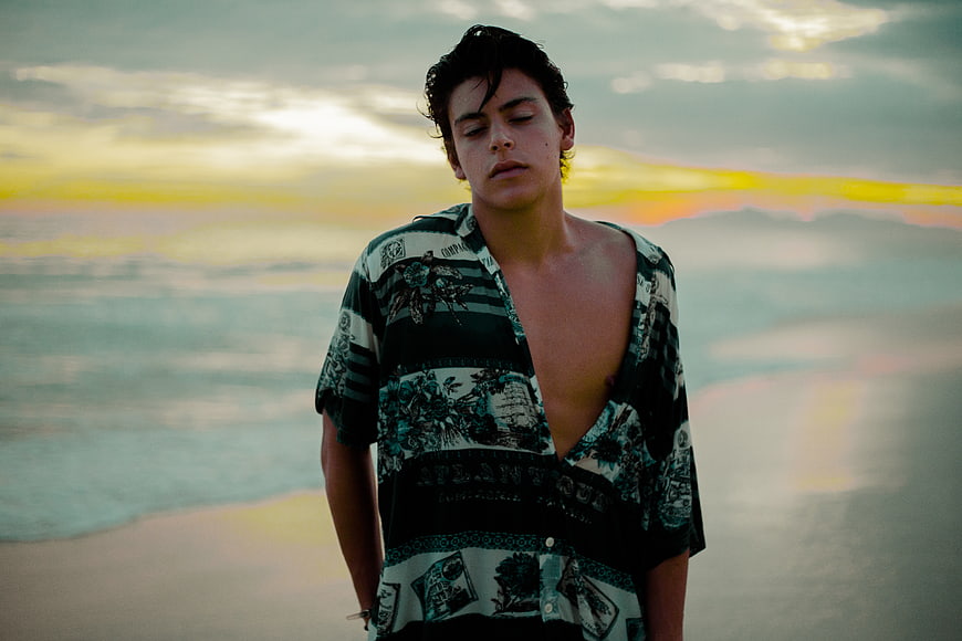 Young man in patterned shirt standing on beach at sunset